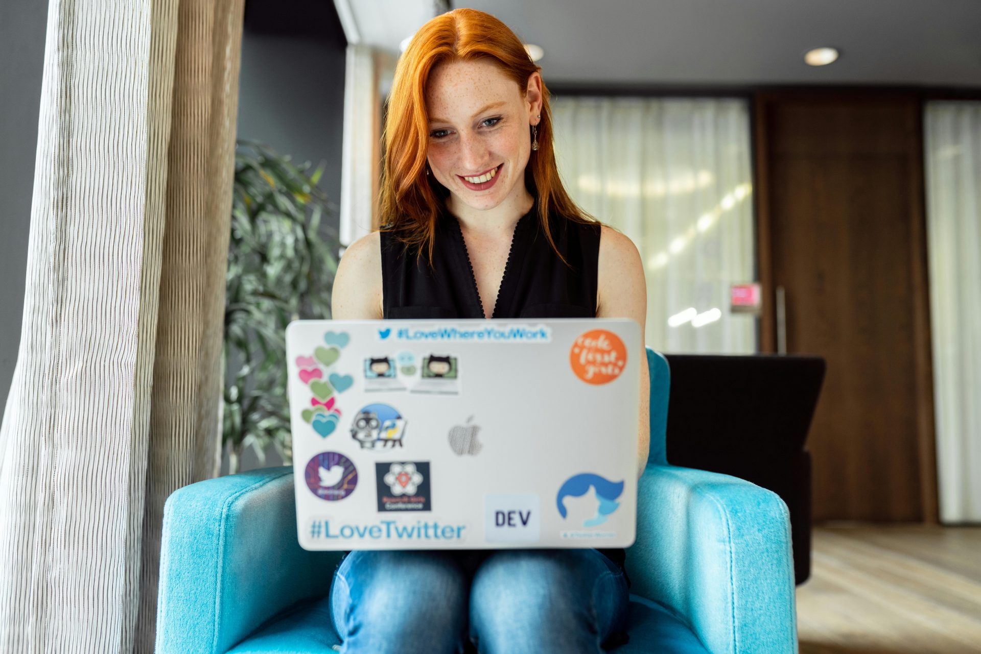 A white woman with red hair smiles while working on a laptop covered in stickers