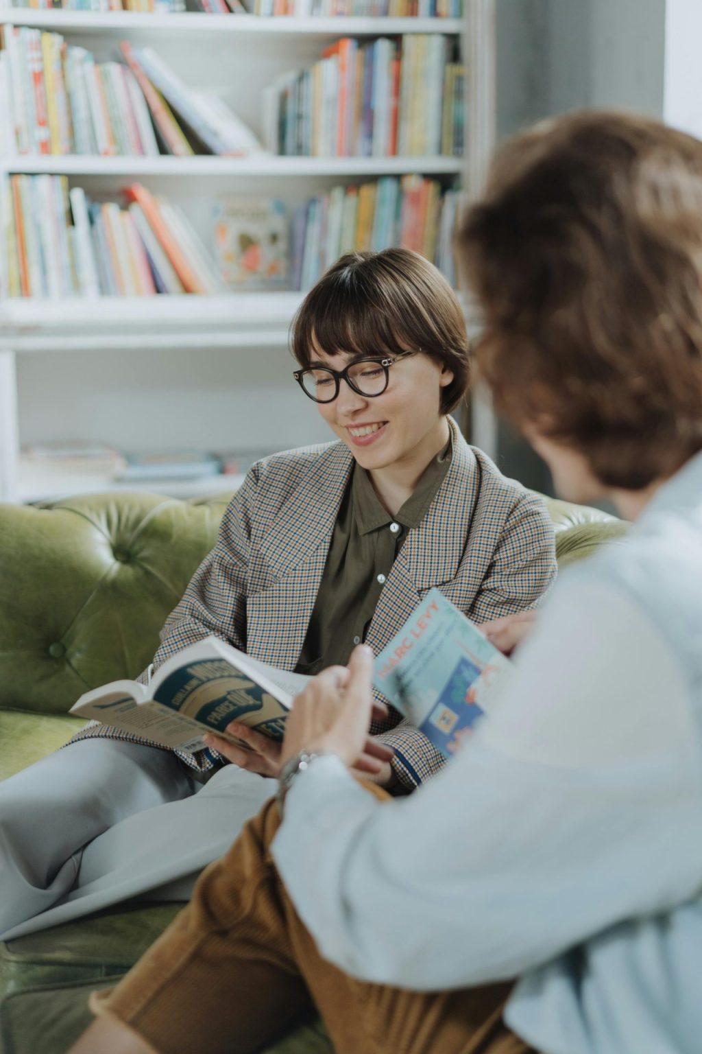 A White woman with short brown hair wears glasses and a brown blazer. She reads a book on a green couch.