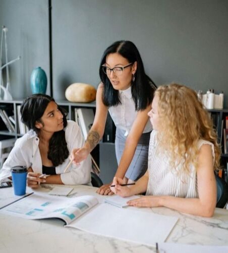 Two women sitting at a table while another woman stands between them, leaning her hands on the table and pointing to a paper in front of them.