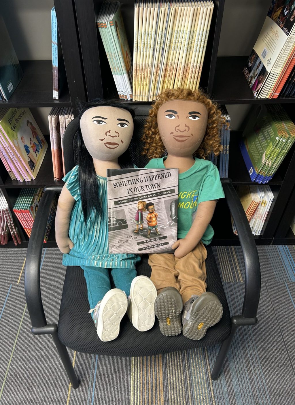 Persona Dolls Leela and Nick sitting in a chair and holding a picture book called Something Happened in Our Town