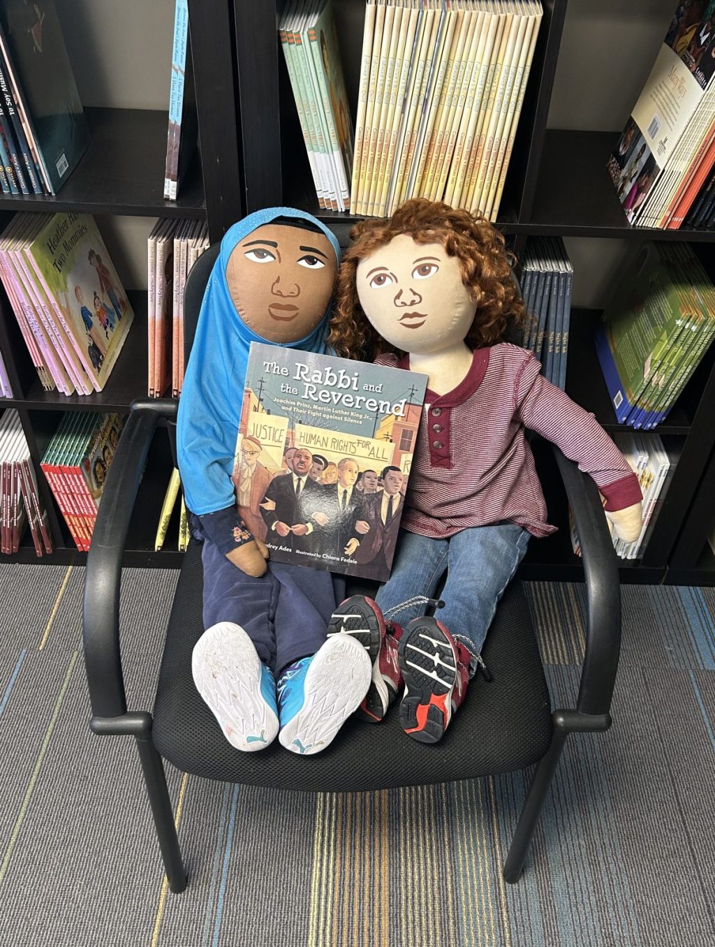 Persona Dolls Rahma and Sam sitting in a chair holding a picture book called The Rabbi and the Reverend