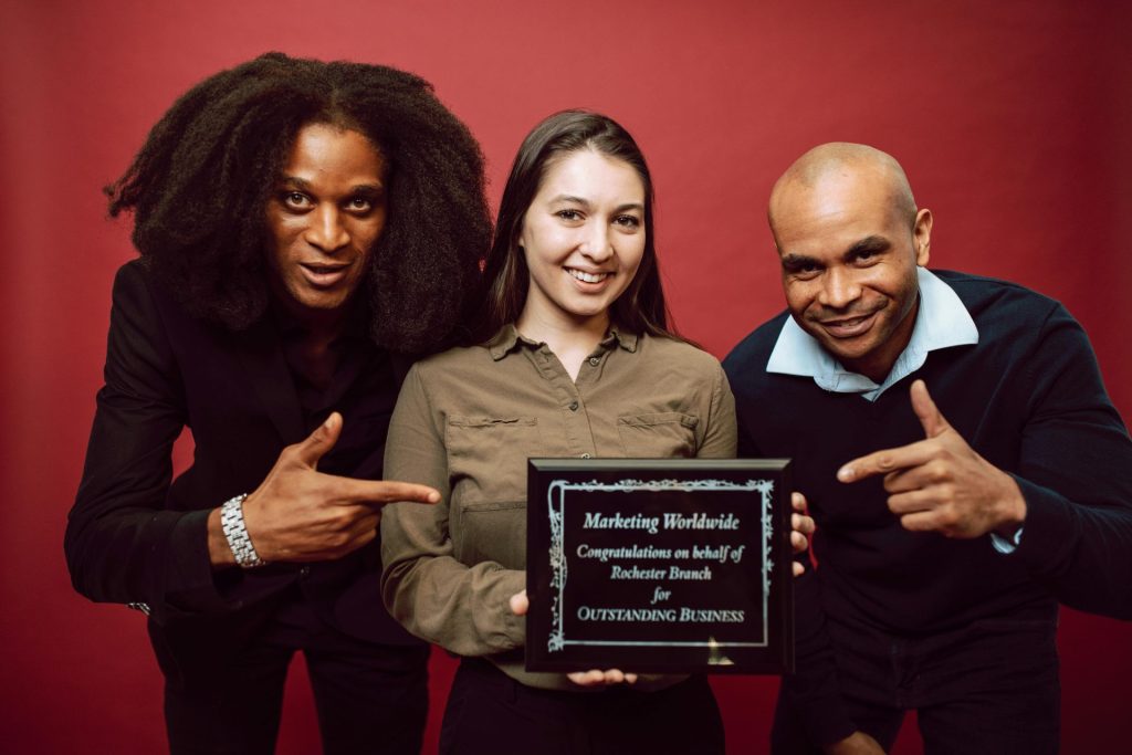 One White woman holds a plaque reading, "Marketing Worldwide: Congratulations on behalf of Rochester Branch for Outstanding Business," while two Black employees smile beside her pointing to the plaque