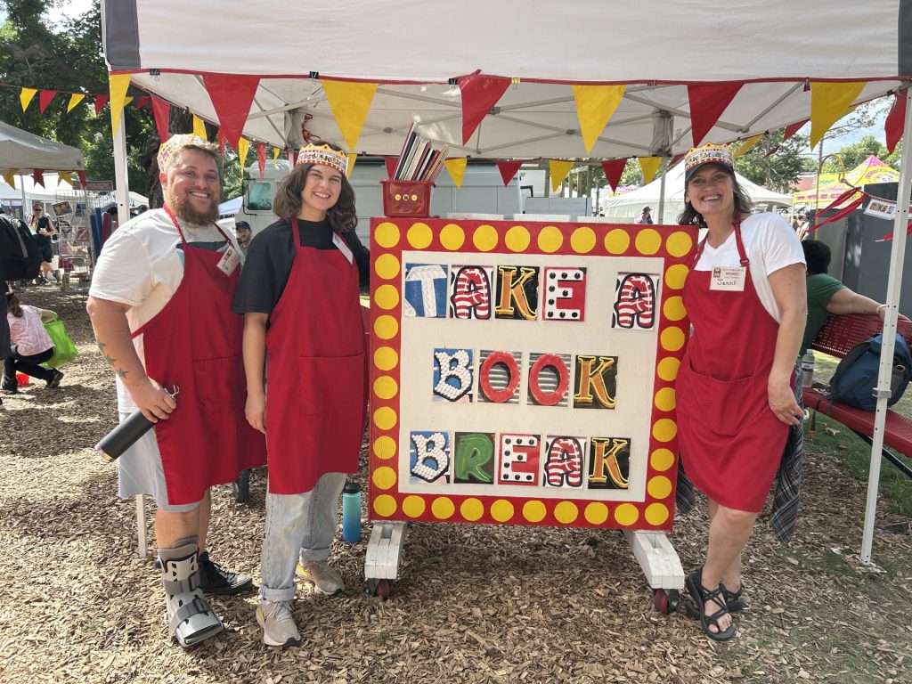 Ryan, Sophie, and Jenni wearing alphabet crowns and red aprons during their Alphabet Forest volunteer shift, posing next to the "Take a Book Break" station.