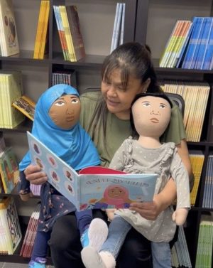 AmazeWorks Persona Doll Artisan sits with two Persona Dolls in her lap, reading a book called "Under My Hijab"